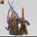 Purple deep dragon miniature viewed from the front, perched among rocky cave stalagmites on a 75mm round base.